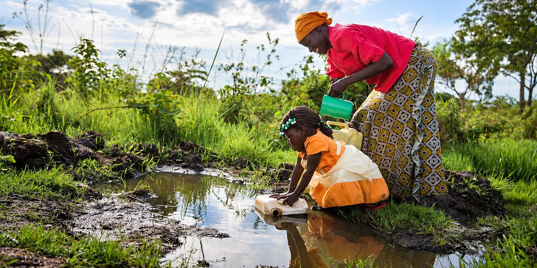 Woman and young girl collecting water at a pond's edge in a rural, tropical region